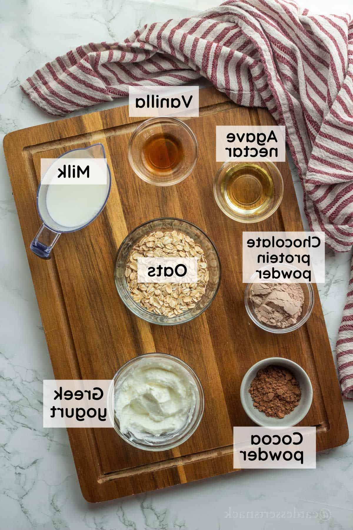 Ingredients for brownie batter overnight oats in glass bowls on wood cutting board with dish towel.