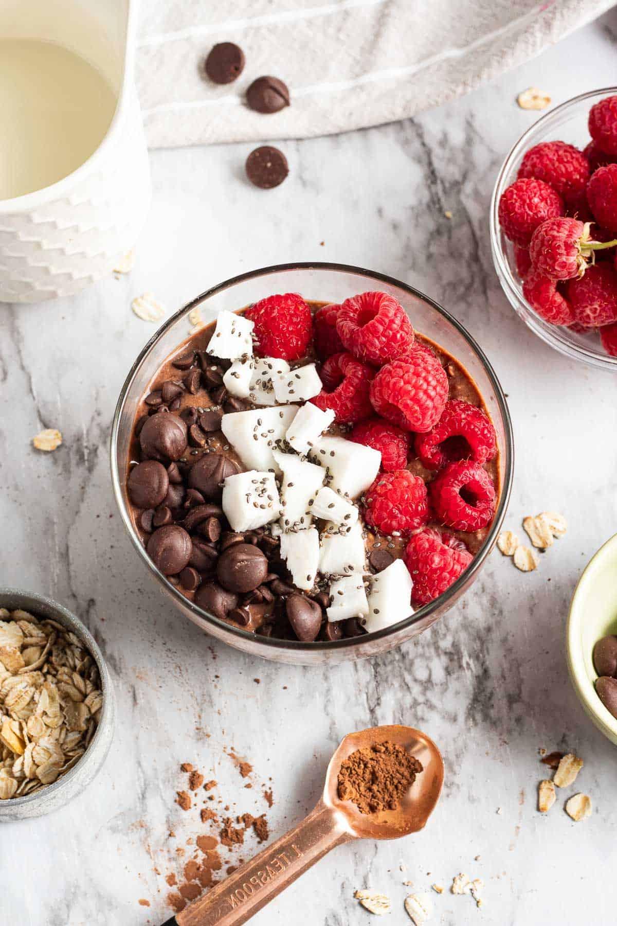 Brownie Batter Overnight Oats with spoonful of cocoa powder and bowl of raspberries nearby.
