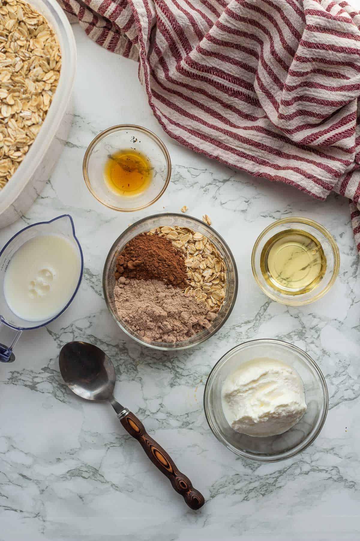 Ingredients for brownie batter overnight oats in glass bowls on marble countertop with dish towel.