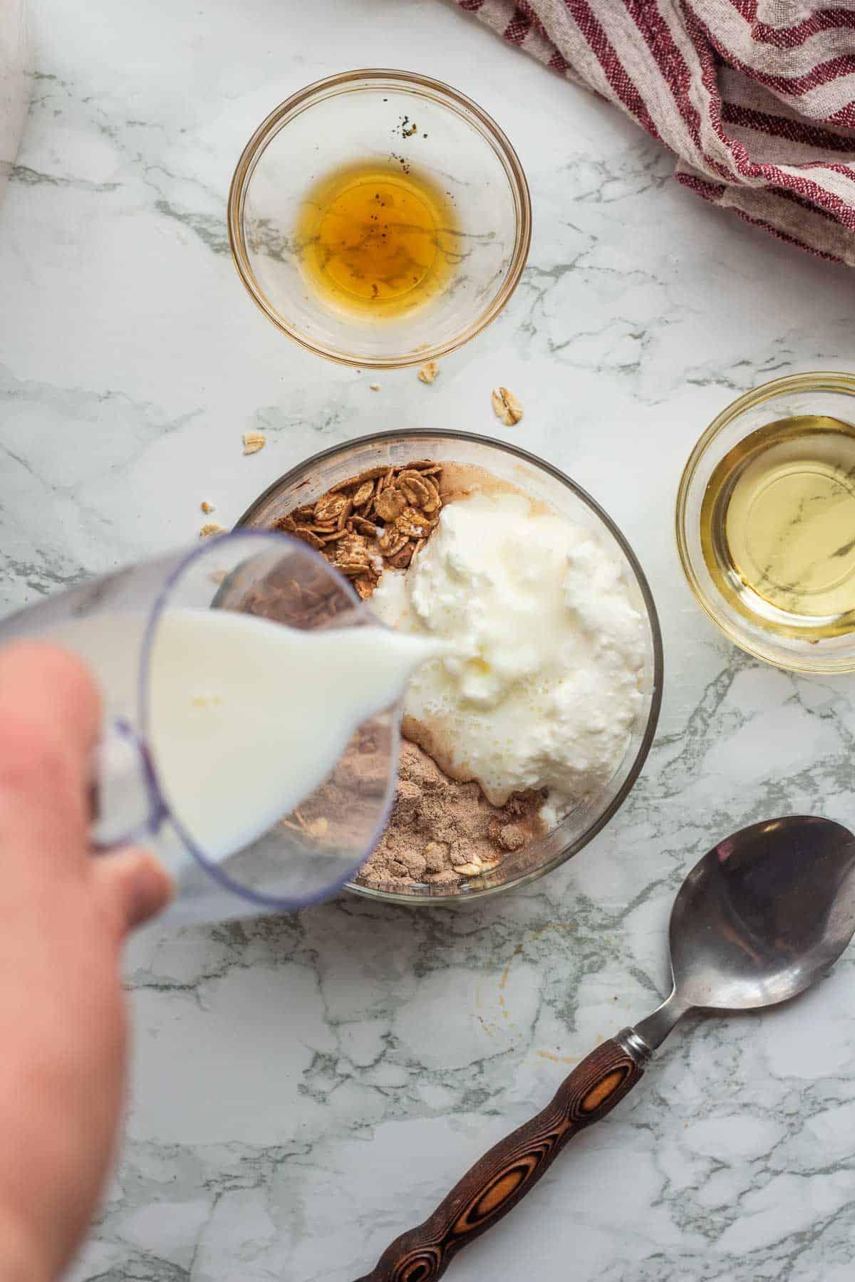 Pouring milk into glass bowl with oats, cocoa powder, protein powder, and greek yogurt.