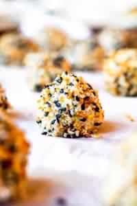 These Mini Everything Bagel Cheese Ball Bites with crackers are the best appetizer recipe for holiday gatherings with friends and family, game days, girls nights, baby showers or birthday parties. These cheese balls are filled with cream cheese, bacon, scallions, and cheese then coated in Everything But the Bagel Seasoning! Perfect to be made ahead of time for easy prep! VIDEO included! #appetizer #recipe #cheeseballs Close up of cheese ball on parchment paper on a cookie sheet.