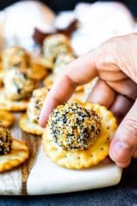 These Mini Everything Bagel Cheese Ball Bites with crackers are the best appetizer recipe for holiday gatherings with friends and family, game days, girls nights, baby showers or birthday parties. These cheese balls are filled with cream cheese, bacon, scallions, and cheese then coated in Everything But the Bagel Seasoning! Perfect to be made ahead of time for easy prep! VIDEO included! #appetizer #recipe #cheeseballs Hand picking up cheese ball bite off of a cutting board.