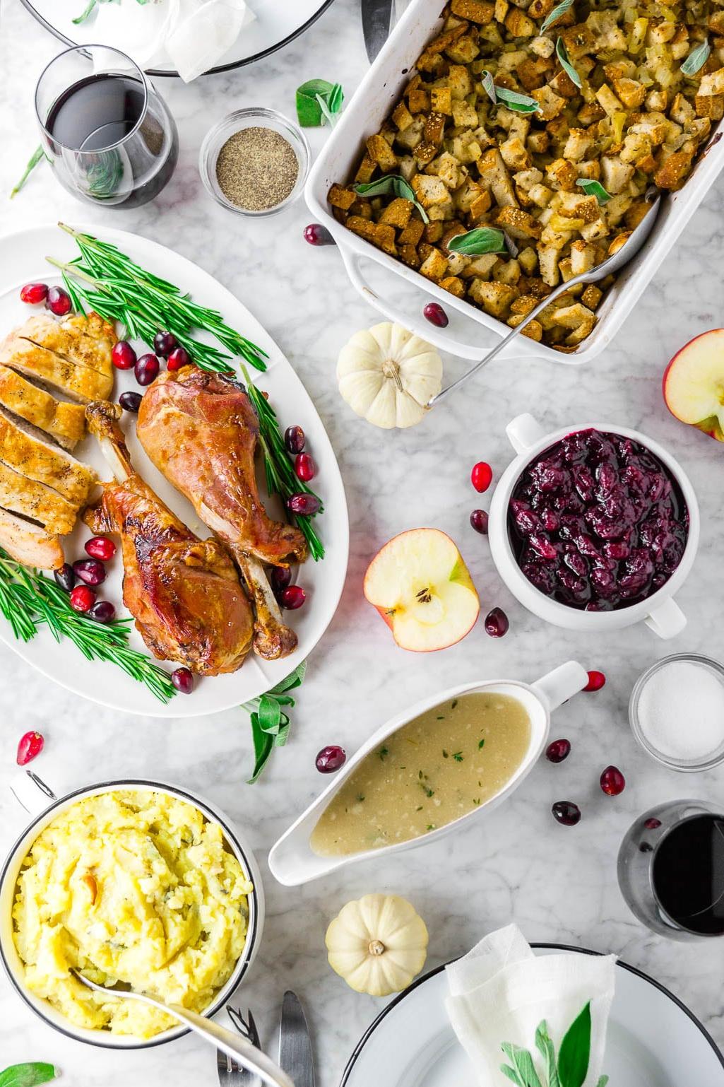 A white marble table with a Thanksgiving feast with gluten-free stuffing, turkey legs with cranberries and rosemary, cranberry sauce, gluten-free grave, a bowl of mashed potatoes, and two glasses of red wine with place settings.