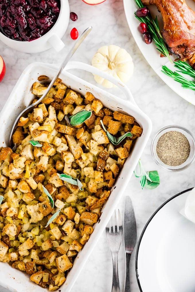 A baking sheet with traditional apple sage gluten-free stuffing with a plate of turkey and a bowl of cranberry sauce and a plate setting ready for Thanksgiving dinner.