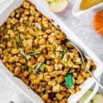 A white baking sheet with classic gluten-free stuffing with celery, onion and apple with a bowl of mashed potatoes and gravy.