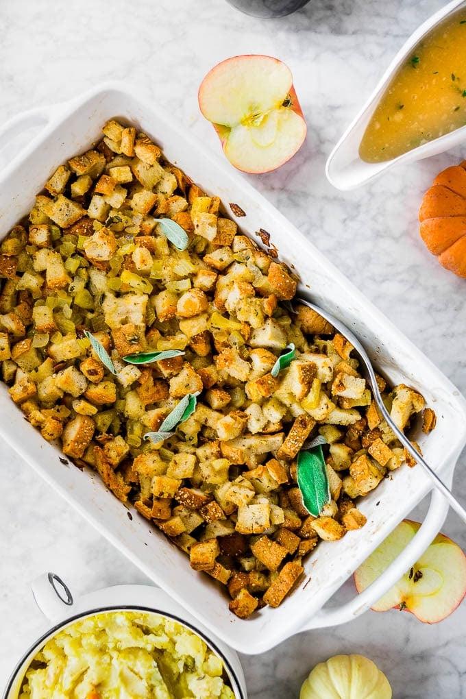 A white baking sheet with classic gluten-free stuffing with celery, onion and apple with a bowl of mashed potatoes and gravy.